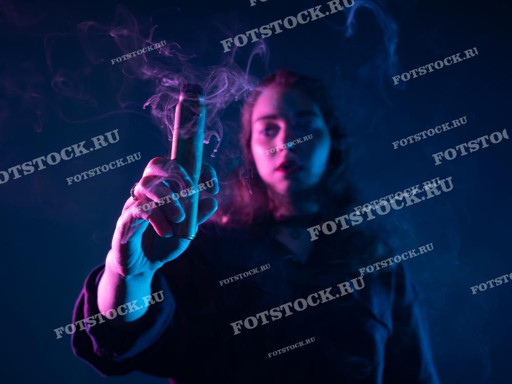 На фотографии изображена молодая женщина, держащая вейп-устройство и окруженная густым цветным дымом. Снимок акцентирует внимание на современной культуре вейпинга и атмосферном освещении.